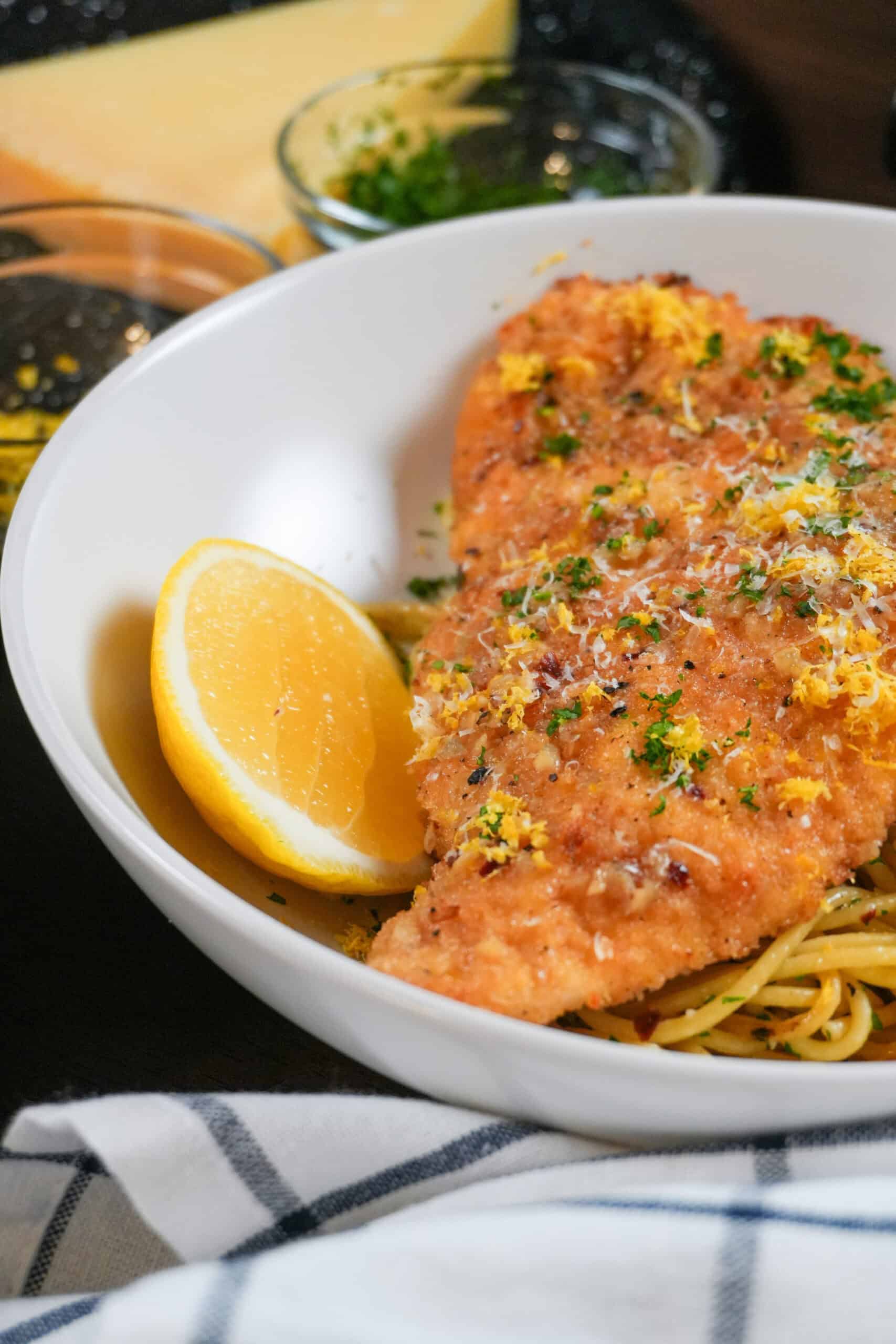 Crispy baked chicken cutlet atop al dente pasta with fresh herbs and lemon wedge garnish.